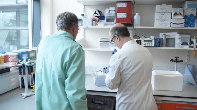 An image of two men wearing lab coats facing away from the camera looking at an experiment in a lab.
