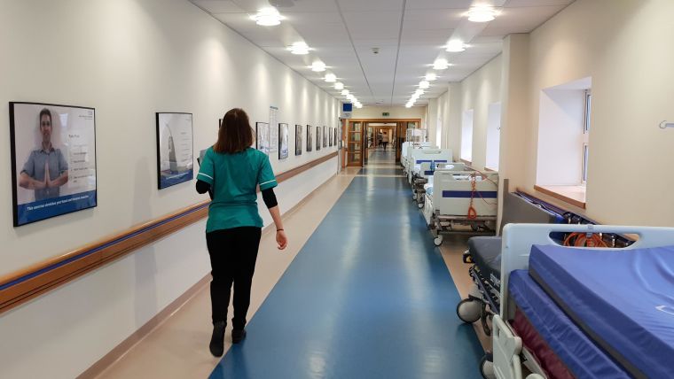 A person walking down an empty hospital hallway with a row of beds to the right.