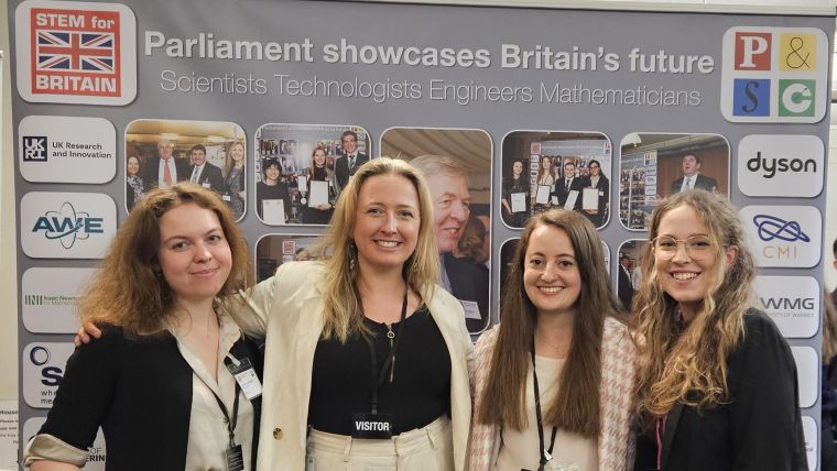 Four women stand smiling in front of a banner, which says "Parliament showcases Britain's future Scientists Technologies Engineers Mathematicians".