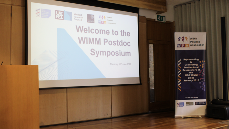 A podium and vertical banner. On the screen reads 'Welcome to the WIMM Postdoc Symposium'.